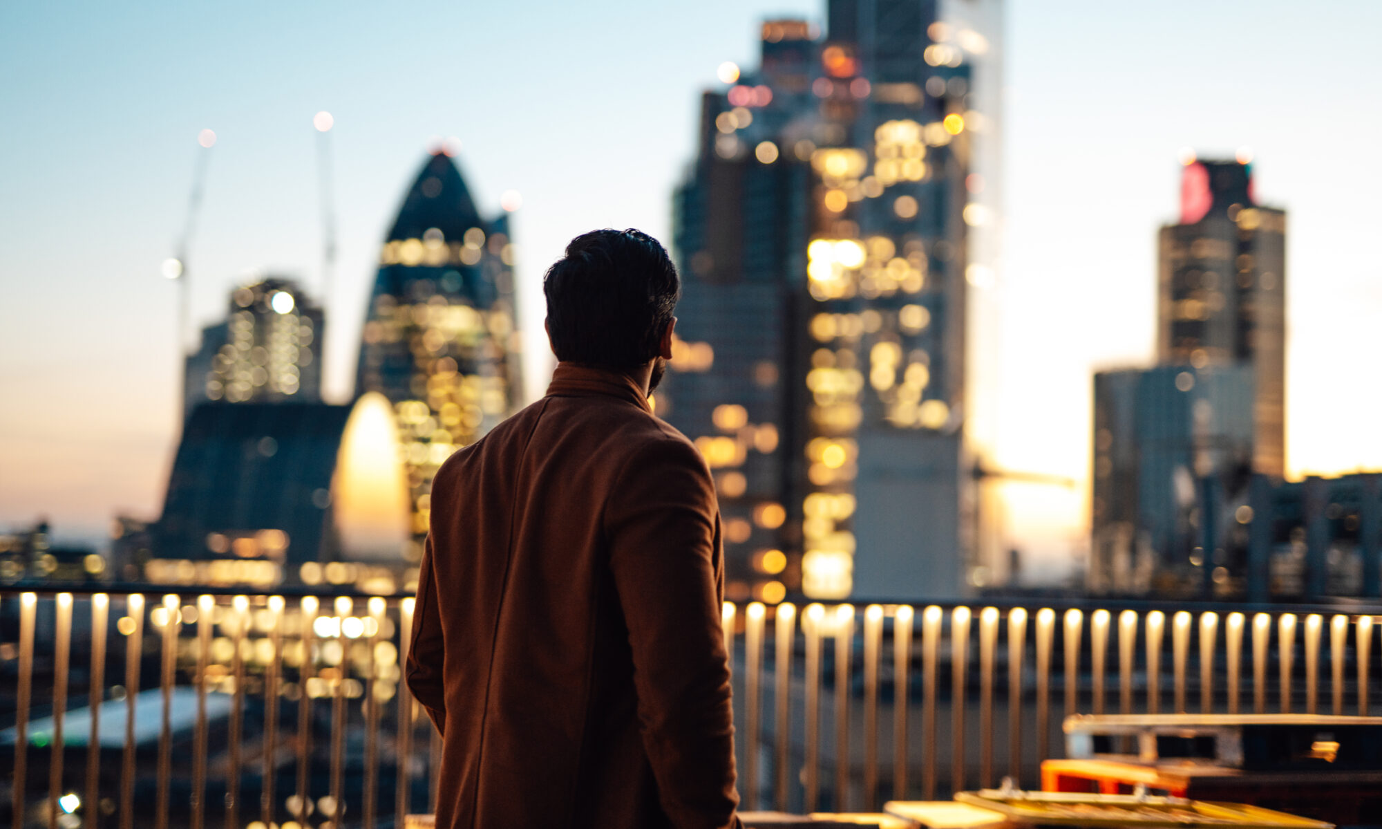 Businessman looking over city at rooftop in high-rise business building against illuminated financial buildings and London cityscape at night.